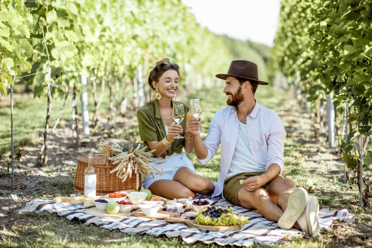 Beautiful couple having romantic breakfast with lots of tasty food and wine, sitting together on the picnic blanket at the vineyard on a sunny morning