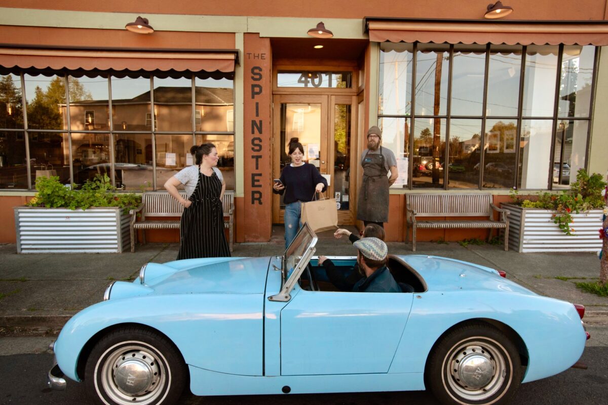 Ela Jean Beedle, center, delivers a meal to Randy Czech, driving, and Erin Mitchell during curbside pickup only service while Jake Ameral, right, and owner Liza Hinman, left, come out to check out their sporty car at The Spinster Sisters restaurant in Santa Rosa on April 2, 2020. (Erik Castro/for Sonoma Magazine)