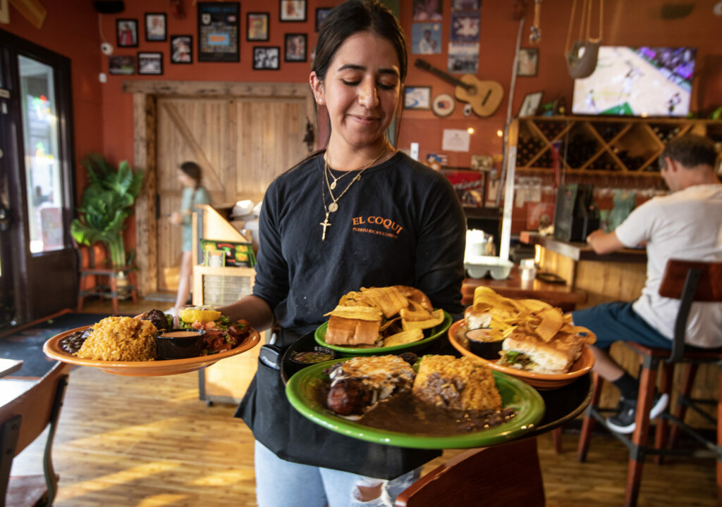 An employee of El Coqui takes plates to guests in Santa Rosa on Wednesday, April 26, 2023. Chef Jackie Roman credits her culinary skills to her mother and grandmother's cooking. From left, bacalaito, a cod fish fritter, served with rice, beans and plantains, rear center, cubano sandwich, pork, ham, Swiss cheese, pickles on a flattened French roll, front center, canoa con carne molida, sweet plantains filled with Angus ground beef, smothered in Monterey Jack cheese, right, a sandwich containing Puerto Rican-seasoned and marinated chicken thighs, served with plantains. (Chad Surmick / The Press Democrat)