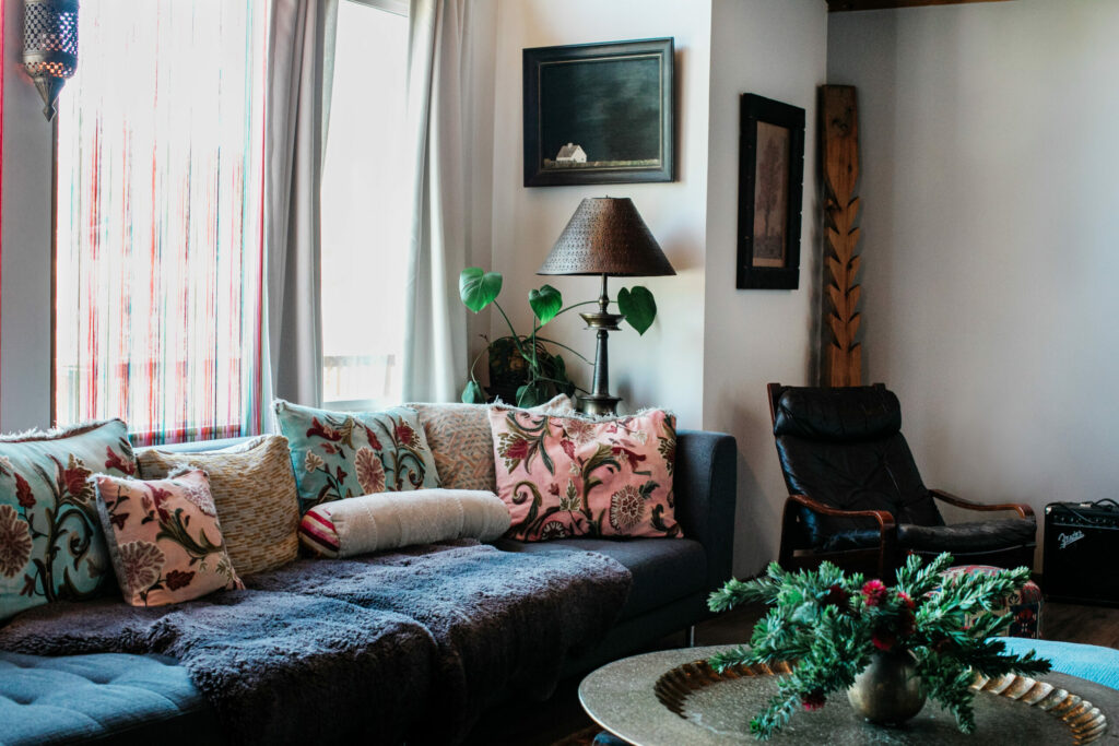 The bright living room, with 1970s-style floor-to-ceiling windows. A quiet farmhouse landscape painted by Moore’s grandmother has a place of honor above the couch. Morgania Moore arranged the holiday flowers and pieced together the sofa cushions from remnants of linen and brocade. (Eileen Roche / Sonoma Magazine)