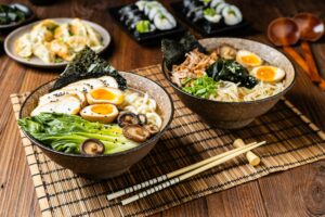 Traditional ramen with jerked pork or chicken. With udon or ramen noodles. Served in classic bowls. Gyoza dumplings and mushrooms in the background. Natural wooden background. gkrphoto/Shutterstock