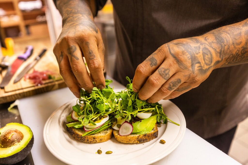 Costeaux Bakery chef Jorge Flores adds pea shoots to top off his Avocado Toast with artisan bread, pepitas, hard-boiled eggs, chile flakes, radishes and pickled red onion at the Healdsburg bakery, Wednesday, June 28, 2023. (John Burgess / The Press Democrat)