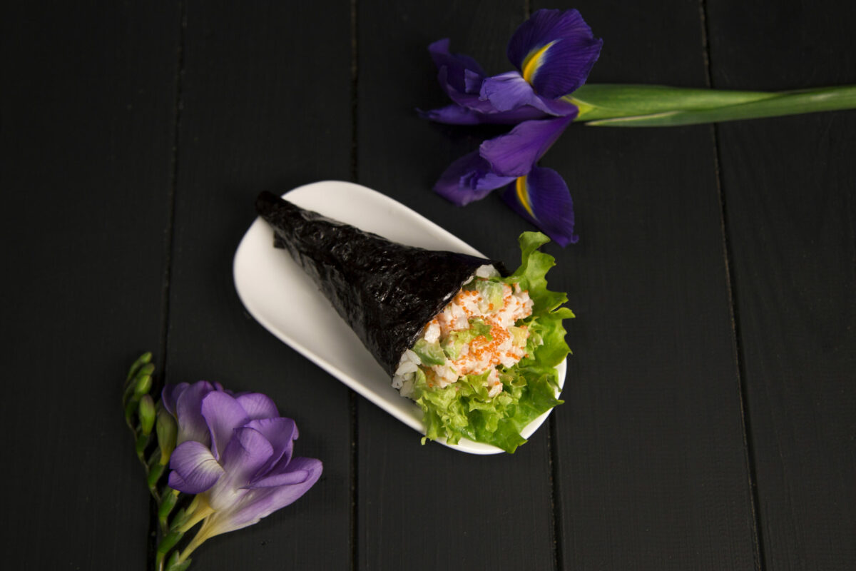 Japanese Sushi Temaki with tiger shrimp, rice, Tobiko roe eggs and salad leaf in nori seaweed on white plate. Iris flower on dark wooden board. Close up of Pan Asian traditional dish. (ArtFood / Getty Images)