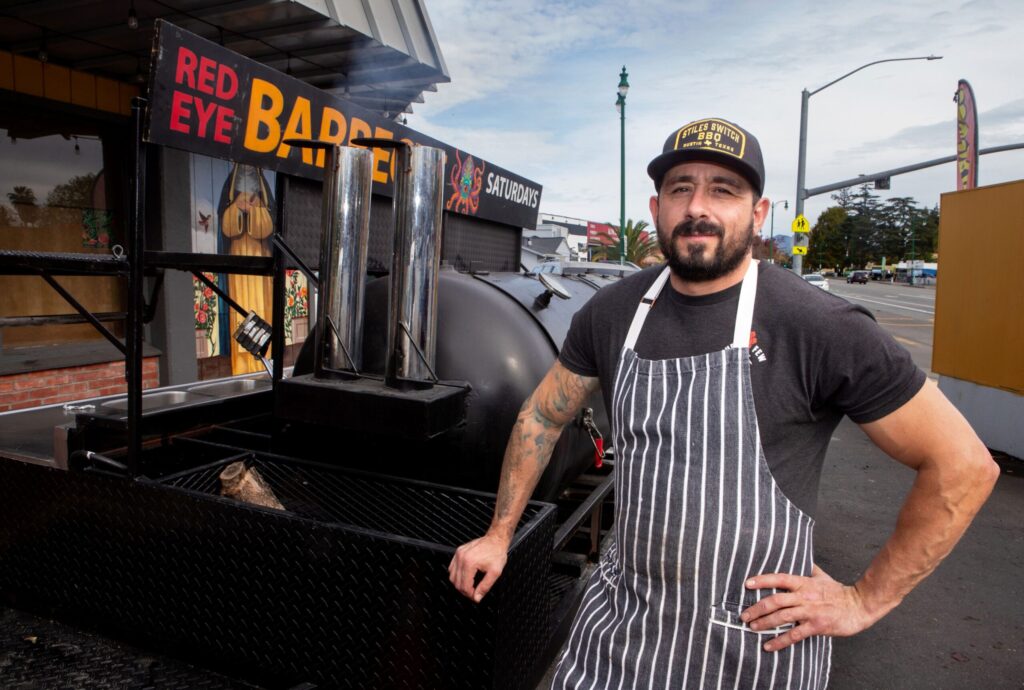 Bob Costarella, owner and pitmaster, stands in front of his smoker at his “Red Eye Barbecue” pop up, held on Saturdays at Sazon Peruvian Cuisine & Deli on Nov. 15, 2025. (Darryl Bush / For The Press Democrat)