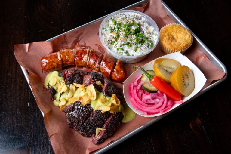 A plate of beef cheek topped with artichoke dill pickle salsa verde sauce including crispy garlic chips, jalapeño cheddar sausage, roasted potato salad, corn bread, and pickled fruits and veggies, are shown at Bob Costarella’s “Red Eye Barbecue” pop-up, held on Saturdays at Sazon Peruvian Cuisine & Deli on Nov. 15, 2025. (Darryl Bush / For The Press Democrat)