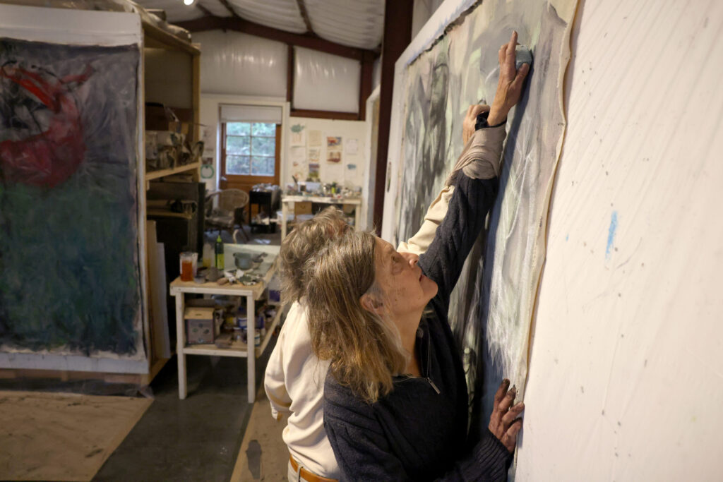 With their arms bound together, artists Pamela Holmes and Winston Gourley draw with bars of pigmented beeswax on a canvas at Stranger Worth studio in Cazadero Thursday, Nov. 20, 2025. (Beth Schlanker / The Press Democrat)
