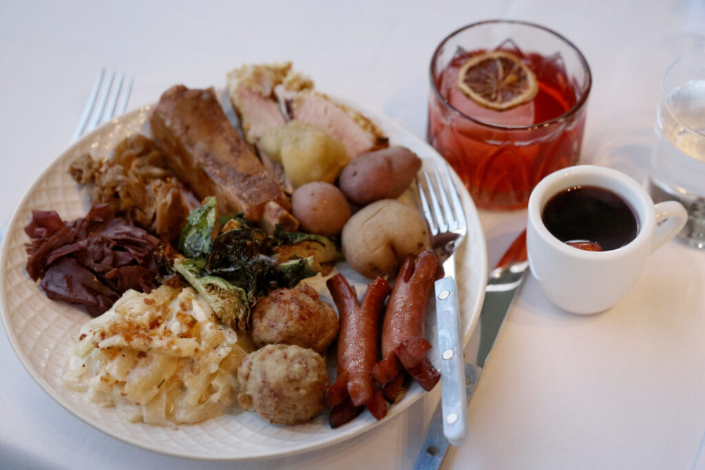(Clockwise from bottom) Swedish meatballs, Jansson Temptation potato and anchovy casserole, fried Brussel sprouts, braised red cabbage with apple, brown cabbage with ginger, ginger glazed spare ribs, sliced Christmas ham topped with applesauce, roasted potatoes, and Frankfurters, with a mug of hot glögg and a Nordgroni cocktail during a holiday julbord at Stockhome in Petaluma Sunday, Dec. 14, 2025. (Beth Schlanker / The Press Democrat)