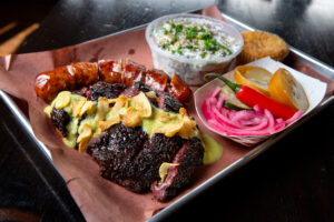 A plate of beef cheek topped with artichoke dill pickle salsa verde sauce including crispy garlic chips, Jalapeño cheddar sausage, roasted potato salad, corn bread, and pickled fruits and veggies, are shown at Bob Costarella’s “Red Eye Barbecue” pop-up, held on Saturdays at Sazon Peruvian Cuisine & Deli on November 15, 2025. (Darryl Bush / For The Press Democrat)