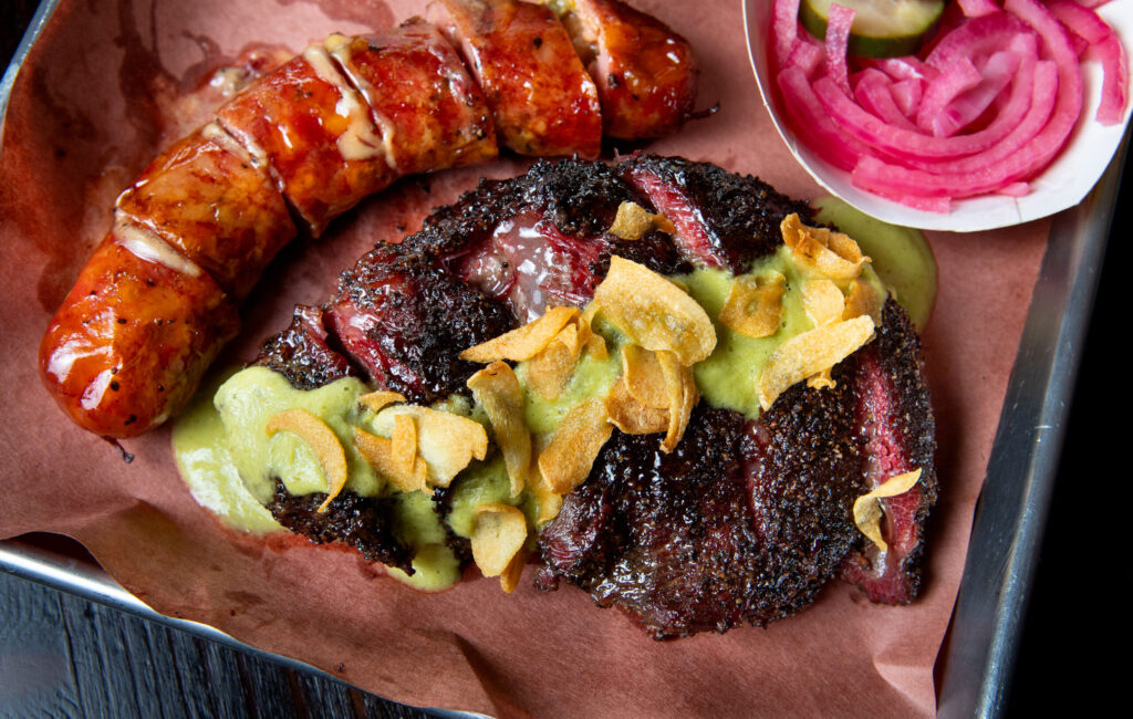 A plate of beef cheek topped with artichoke dill pickle salsa verde sauce including crispy garlic chips, Jalapeño cheddar sausage, roasted potato salad, and pickled fruits and veggies, are shown at Bob Costarella’s “Red Eye Barbecue” pop-up, held on Saturdays at Sazon Peruvian Cuisine & Deli on November 15, 2025. (Darryl Bush / For The Press Democrat)