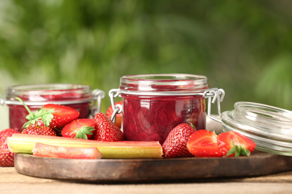Tasty rhubarb jam in jars, stems and strawberries on wooden table against blurred background. (Liudmila Chernetska / Getty Images)