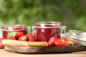 Tasty rhubarb jam in jars, stems and strawberries on wooden table against blurred background. (Liudmila Chernetska / Getty Images)