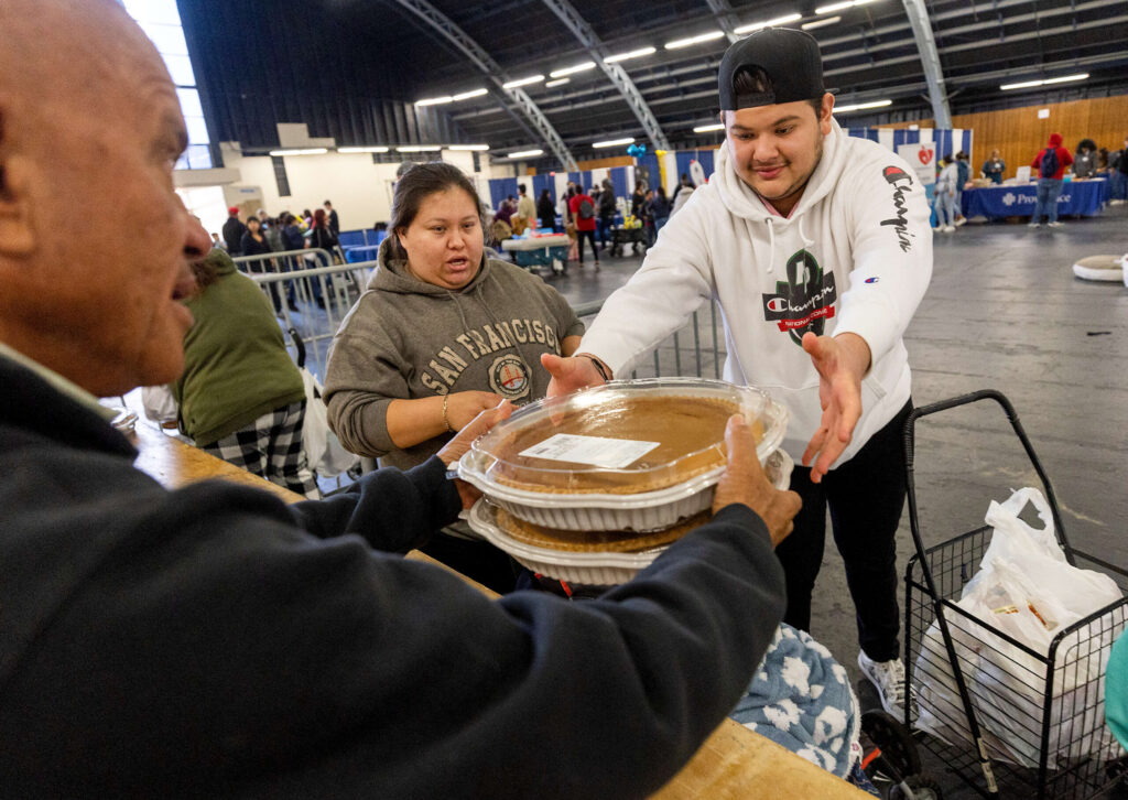 Volunteer Steffan O’Neill hands out pumpkin pies, turkeys and all the fixings for a holiday meal to Valentina Medina, left, and Eduardo Alvarez at the Redwood Gospel Mission Thanksgiving Banquet at the Sonoma County Fairgrounds in Santa Rosa, Wednesday, Nov. 22, 2023. (John Burgess / The Press Democrat)