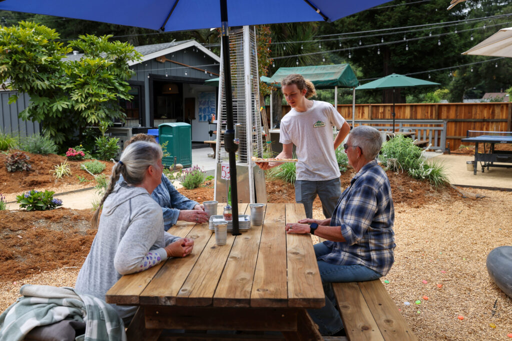 Rio McFarland brings out a pizza to Kim Adams, left, Patti Mauerman, and Leslie Moffatt at Wonderland Pizzeria in Monte Rio on Wednesday, June 17, 2026. (Christopher Chung/The Press Democrat)