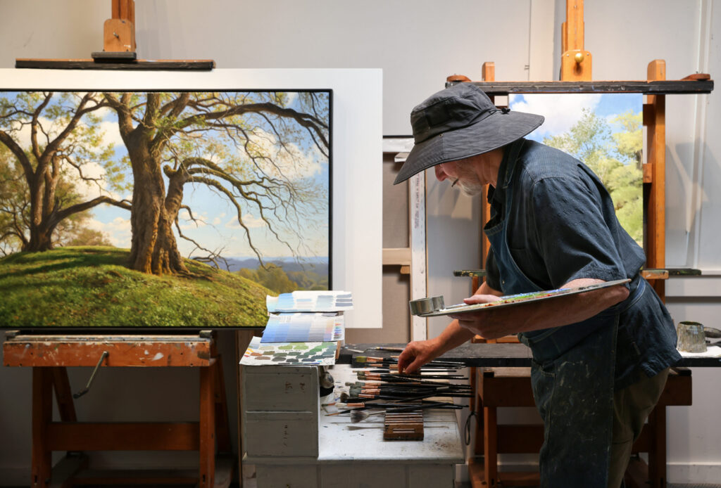 Christopher Leith Evans picks out a brush while working on a landscape at his home studio in Healdsburg on Monday, May 11, 2026. The painting on the left is a scene from Pepperwood Preserve. (Christopher Chung/The Press Democrat)
